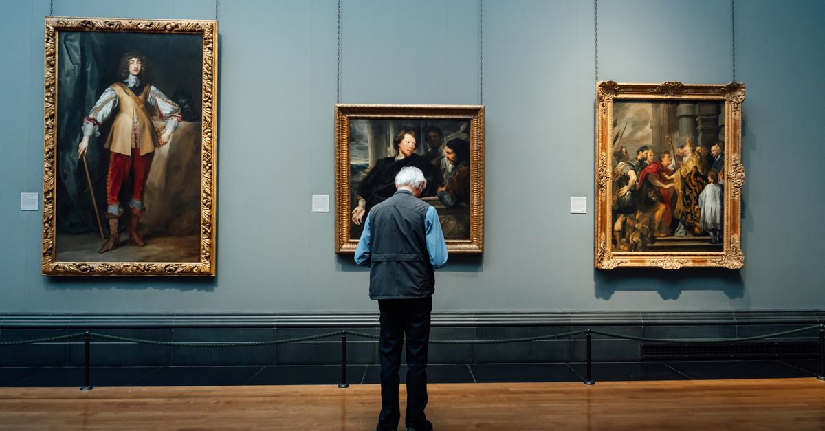 An elderly man admires classic paintings at an art gallery in England. Indoor lighting enhances the historic ambiance.